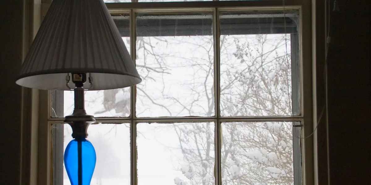 Tischlampe mit weißem Faltenschirm und einem tropfenförmigen Unterteil aus blauem Glas vor einem Sprossenfenster, draußen verschneite Äste